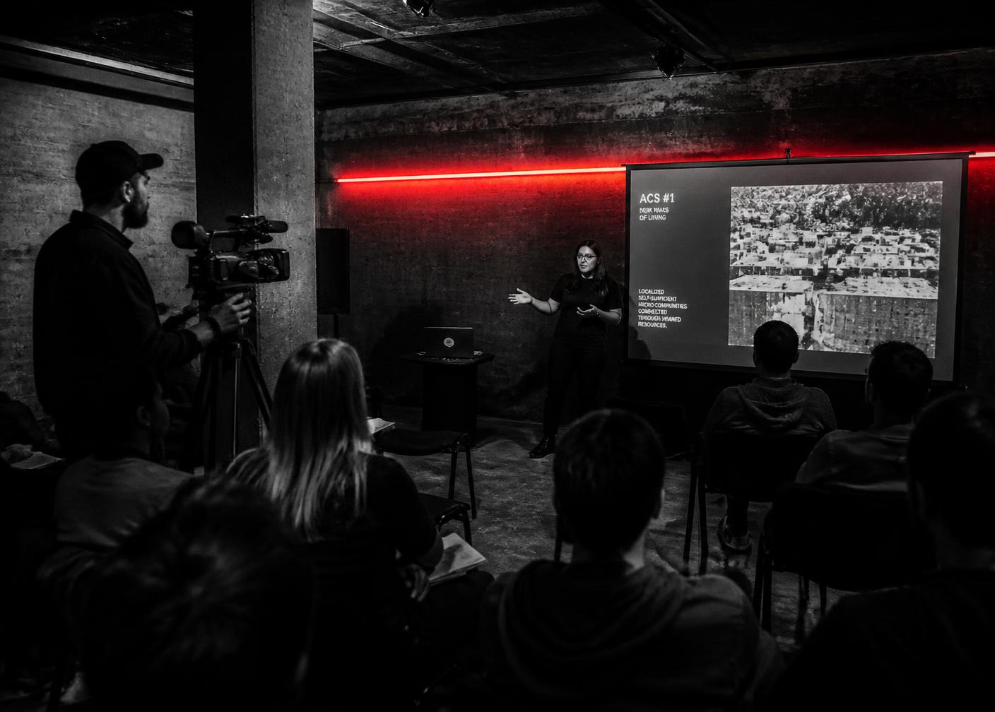 A woman specialist speaking to a seated audience in a dark concrete room while being filmed
